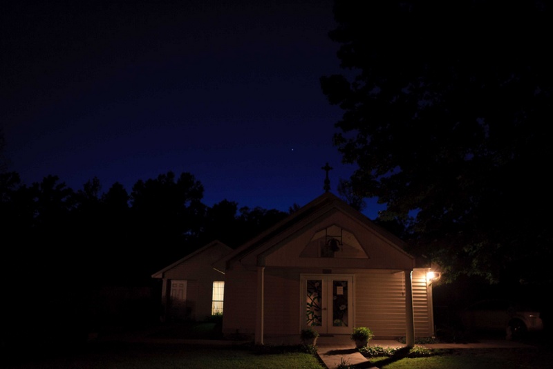A small chapel building at night lit by an outdoor lamp, with a cross on the roof against a dark blue sky